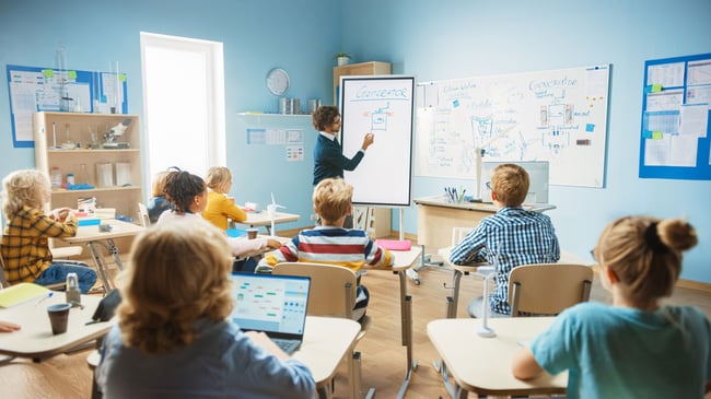 Teacher in front of classroom pointing to a white board.