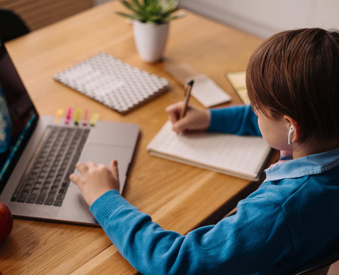 Young male student writing on a notepad while studying with a laptop on a wooden table