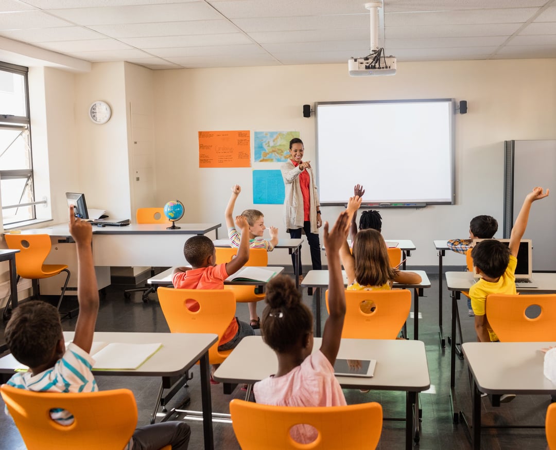 students in a bright classroom raising their hands while a teacher stands at the front near an interactive whiteboard